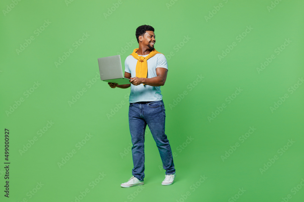 Full body young man of African American ethnicity 20s wear blue t-shirt hold use work on laptop pc computer look aside on workspace isolated on plain green background studio. People lifestyle concept. Full body young man of African American ethnicity 20s wear blue t-shirt hold use work on laptop pc computer look aside on workspace isolated on plain green background studio. People lifestyle concept.