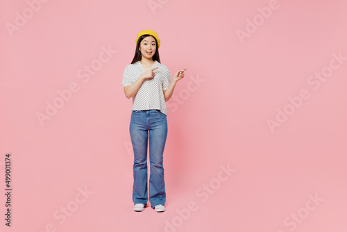 Full body young woman of Asian ethnicity wear white polka dot t-shirt yellow beret point index finger aside on workspace area mock up isolated on plain pastel pink background People lifestyle concept