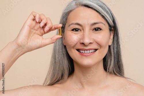 Closeup cropped mature middle-aged caucasian naked shirtless woman holding a pill for beautification, losing weight, healthy dieting additive, health care, painkiller isolated over beige background