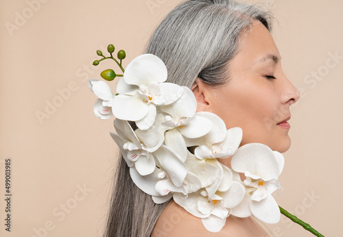Skin and hair care. Anti-age anti-wrinkle beauty products cosmetics. Beautification and rejuvenation. Closeup photo of mature middle-aged woman holding orchid flower on her face with eyes closed photo