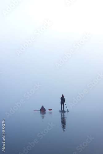 Vertical image of two women paddling kayak and stand up paddle board (SUP) at autumn misty river at foggy autumn morning
