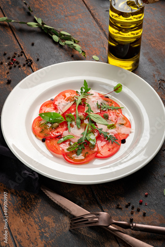 Light Salad with thinly sliced tomatoes, arugula, parmesan, capers and balsamic sauce on white plate. Vegetarian tomato salad on wooden table