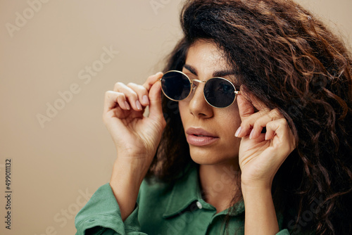 EYEWEAR CONCEPT. Pretty stylish tanned curly Latin female in casual things wear sunglasses posing isolated over pastel beige background look aside. Copy space Mockup Banner. Fashion closeup offer