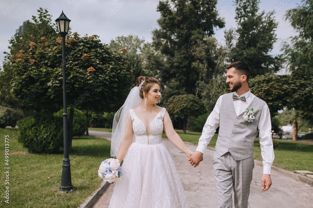 A young, bearded groom in a gray suit and a beautiful, sweet bride in a white dress are walking along the road in nature in the park, garden, holding hands. Wedding portrait, photo of newlyweds.