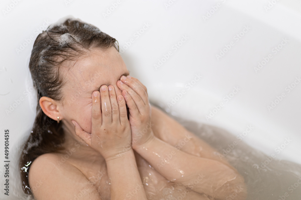 Little, beautiful girl, baby bathes, sitting in a white bath with foam and hiding his hands in his face Little, beautiful girl, baby bathes, sitting in a white bath with foam and hiding his hands in his face
