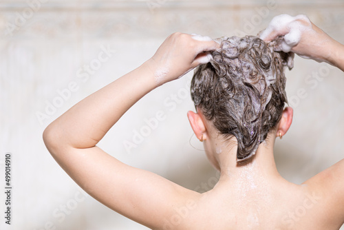 Little, beautiful girl, baby bathes sitting in a white bath with soap suds, shampoo and washes her head with hair. With your back to the camera