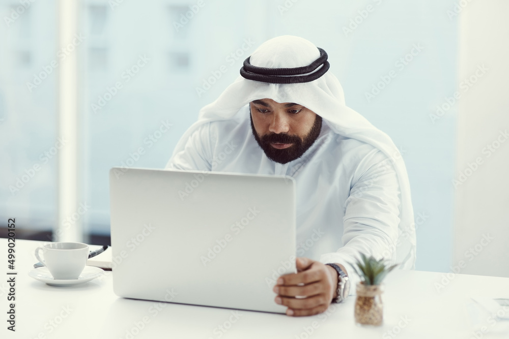 Focused on the task at hand. Cropped shot of a young businessman dressed in Islamic traditional clothing working on his laptop while sitting in the office. Focused on the task at hand. Cropped shot of a young businessman dressed in Islamic traditional clothing working on his laptop while sitting in the office.