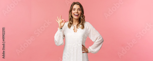 Portrait of carefree good-looking blond girl, wear white dress, guarantee you will like this special spring offer, show okay sign, agree or accept, smiling and nod in approval, pink background