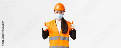 Covid-19 safety protocol at enterpise, construction and preventing virus concept. Confident female asian industrial worker, engineer in face mask and helmet showing thumbs-up, all good photo