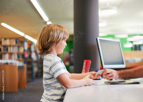 Portrait of cute little boy taking books in library, giving  membership card to librarian