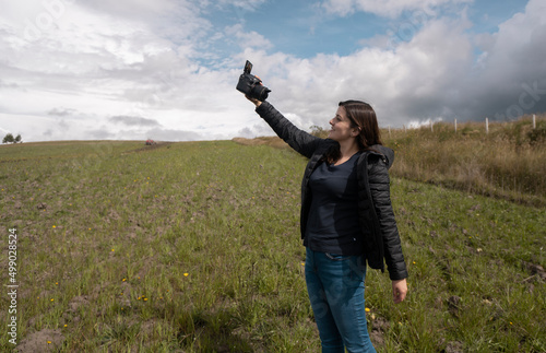 Beautiful hispanic woman blogger filming herself with her camera in her hand happy posing in the middle of the field