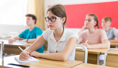 Attractive smart teen girl in glasses studying in classroom, listening to lecturer and writing in notebook