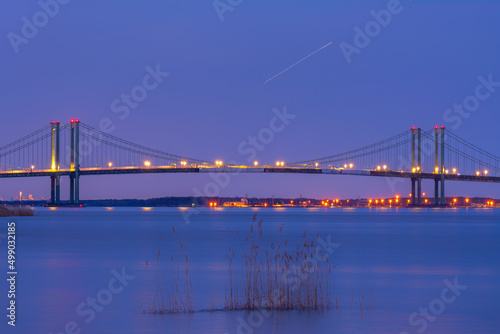 Delaware Memorial Bridge with lights on at dawn colored sky and smooth blue water of Delaware river and reedy grass in foreground.