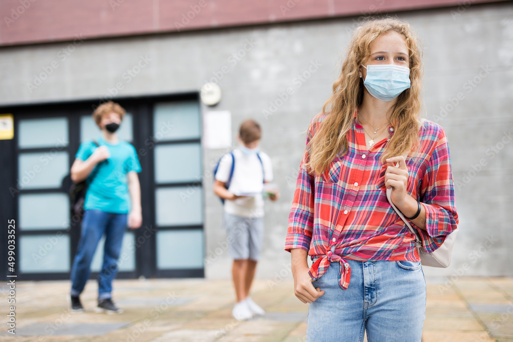 Young girl in face mask with backpack standing near entrance to school building. Teenagers leaving school in background..