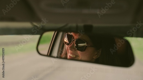 Beautiful young woman traveling by car, smiling, rejoicing. Young woman driver in sunglasses in rearview mirror reflection. Girl driver is driving her car. Weekends, car, adventure, road and sports.