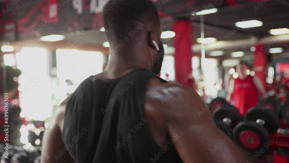 Back view of african american man bodybuilder with wireless headphones performing Side raise exercise with dumbbells at gym Back view of african american man bodybuilder with wireless headphones performing Side raise exercise with dumbbells at gym