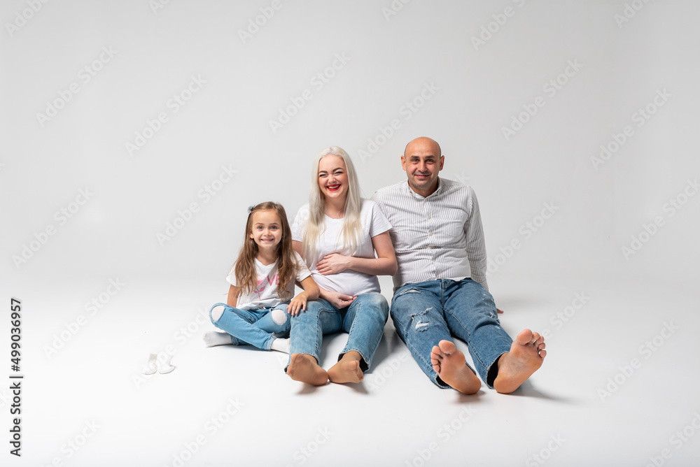Happy family of three sitting on the floor, light background. Laughs: mother, father and little boy. Mom is pregnant. Good news, pregnant, bare feet to us Happy family of three sitting on the floor, light background. Laughs: mother, father and little boy. Mom is pregnant. Good news, pregnant, bare feet to us
