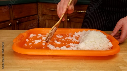 Home cooking - using spatula to spread cooked sushi rice onto orange plastic tray preparing for cooling.