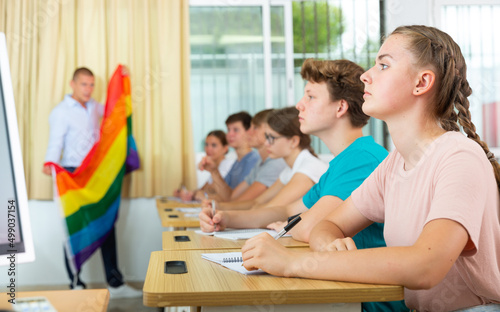 Pupils sitting in class and listening carefully to male teacher. He holding LGBT pride flag in hands and talking about sex minorities.