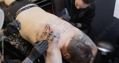 Two tattoo artists working simultaniously on a male back. The closest artist wipes off ink.
4K static shot. photo