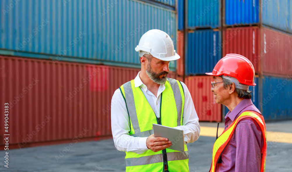 Caucasian and Asian workers teamwork men wear safety clothing. Work clothes with hard hats and use a tablet to inspect containers Caucasian and Asian workers teamwork men wear safety clothing. Work clothes with hard hats and use a tablet to inspect containers
