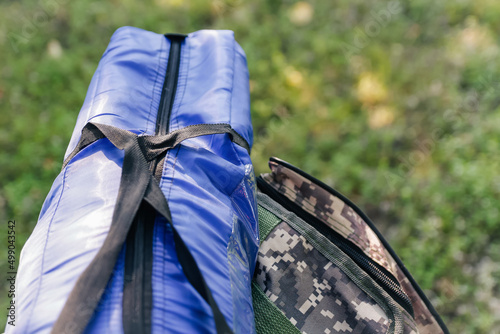 Defocus military backpack and blue tent or sleeping bag. Army bag on green grass background near tree. Military camouflage army rucksack. Tourist summer hiker. Summer. Close-up. Outdoor. Out of focus