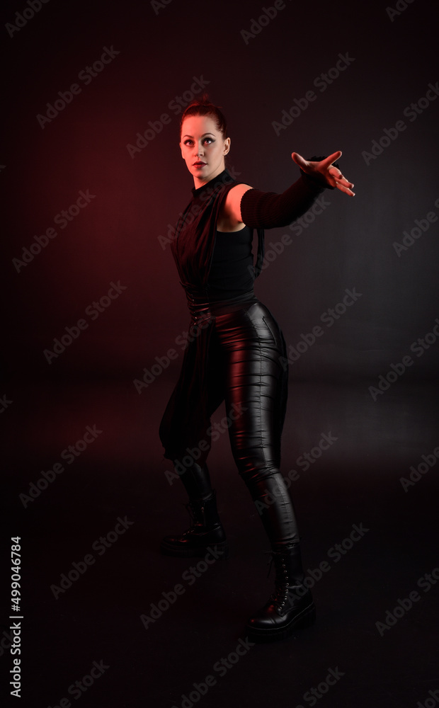Full length portrait of pretty redhead female model wearing black futuristic scifi leather cloak costume. Standing pose on dark studio background with shadow rim moody lighting. Full length portrait of pretty redhead female model wearing black futuristic scifi leather cloak costume. Standing pose on dark studio background with shadow rim moody lighting.