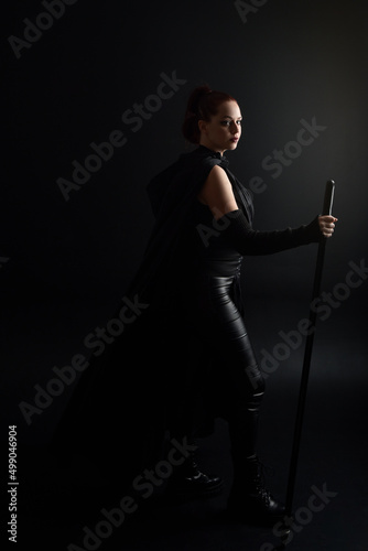 Full length portrait of pretty redhead female model wearing black futuristic scifi leather cloak costume. Standing pose on dark studio background with shadow rim moody lighting.