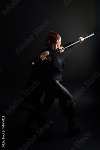 Full length portrait of pretty redhead female model wearing black futuristic scifi leather cloak costume. Standing pose on dark studio background with shadow rim moody lighting.