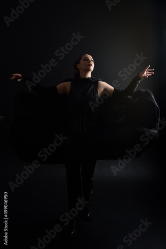 Full length portrait of pretty redhead female model wearing black futuristic scifi leather cloak costume. Standing pose on dark studio background with shadow rim moody lighting.