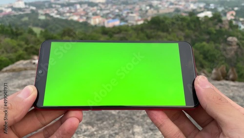 Using phone with green screen chroma key horizontal landscape close up man hand holding smartphone. Monkey relaxing on top of rock mountain, forest, cityscape panoramic view in background