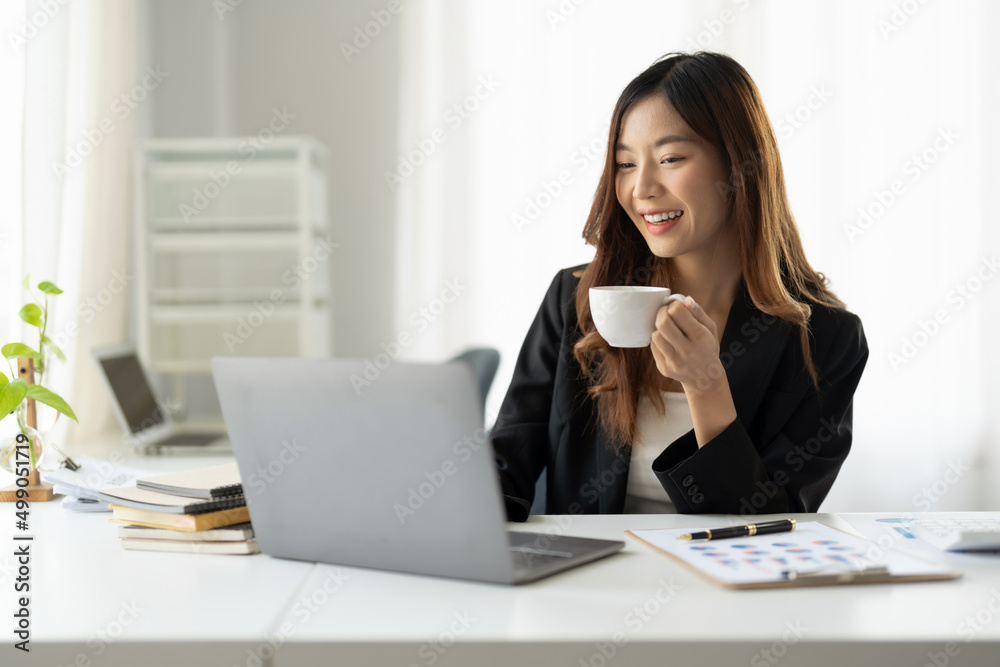 Happy charming asian businesswoman holding a cup of coffee while working on laptop in morning Happy charming asian businesswoman holding a cup of coffee while working on laptop in morning