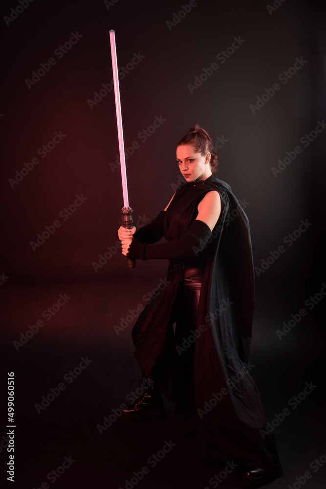 Full length portrait of pretty redhead female model wearing black futuristic scifi leather cloak costume. Standing pose holding lightsaber on dark studio background with shadow moody lighting. Full length portrait of pretty redhead female model wearing black futuristic scifi leather cloak costume. Standing pose holding lightsaber on dark studio background with shadow moody lighting.