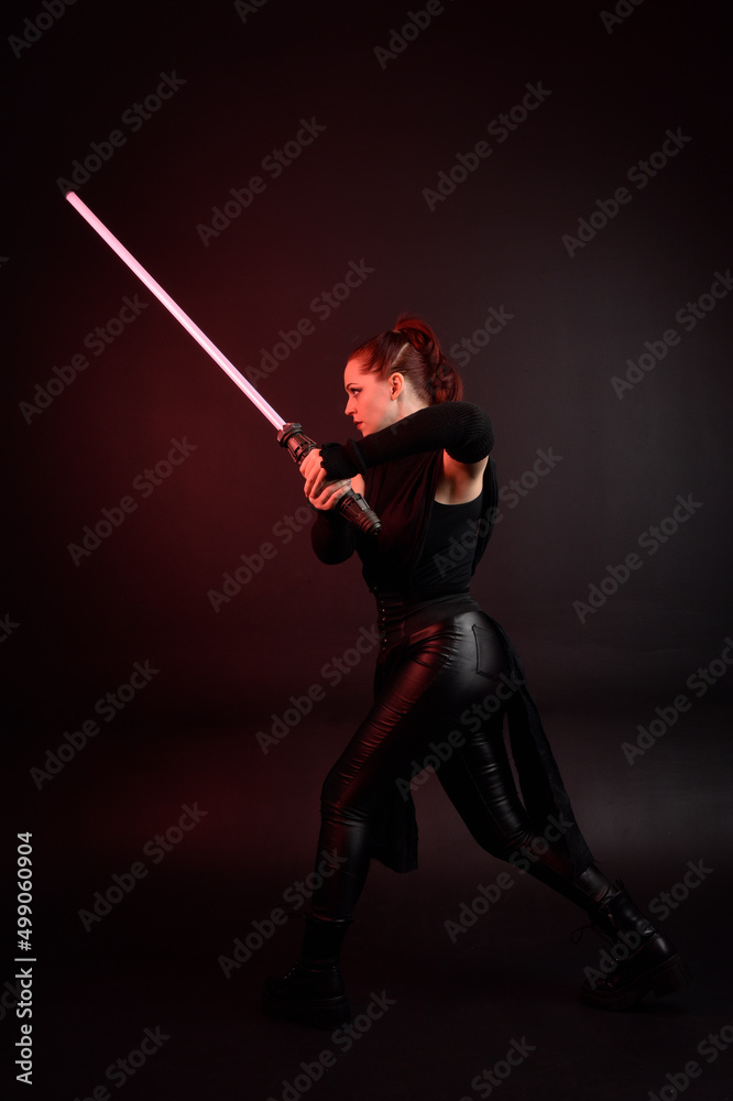 Full length portrait of pretty redhead female model wearing black futuristic scifi leather cloak costume. Standing pose holding lightsaber on dark studio background with shadow moody lighting. Full length portrait of pretty redhead female model wearing black futuristic scifi leather cloak costume. Standing pose holding lightsaber on dark studio background with shadow moody lighting.