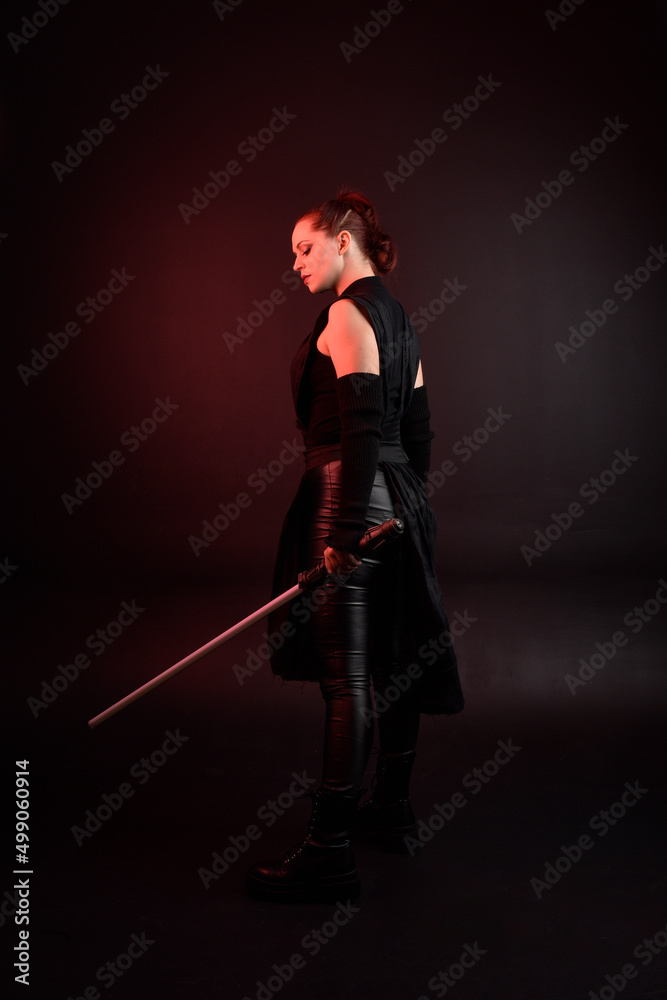 Full length portrait of pretty redhead female model wearing black futuristic scifi leather cloak costume. Standing pose holding lightsaber on dark studio background with shadow moody lighting. Full length portrait of pretty redhead female model wearing black futuristic scifi leather cloak costume. Standing pose holding lightsaber on dark studio background with shadow moody lighting.