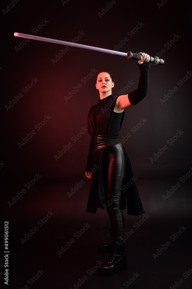 Full length portrait of pretty redhead female model wearing black futuristic scifi leather cloak costume. Standing pose holding lightsaber on dark studio background with shadow moody lighting. Full length portrait of pretty redhead female model wearing black futuristic scifi leather cloak costume. Standing pose holding lightsaber on dark studio background with shadow moody lighting.