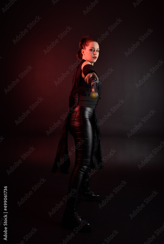 Full length portrait of pretty redhead female model wearing black futuristic scifi leather cloak costume. Standing pose  holding lightsaber on dark studio background with shadow moody lighting.