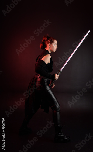 Full length portrait of pretty redhead female model wearing black futuristic scifi leather cloak costume. Standing pose holding lightsaber on dark studio background with shadow moody lighting.