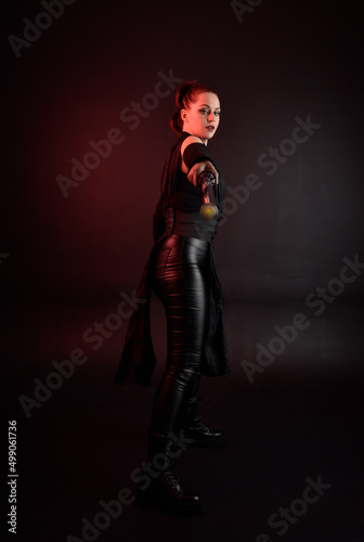 Full length portrait of pretty redhead female model wearing black futuristic scifi leather cloak costume. Standing pose  holding lightsaber on dark studio background with shadow moody lighting. © faestock