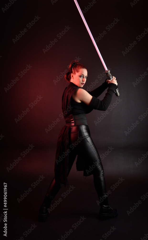Full length portrait of pretty redhead female model wearing black futuristic scifi leather cloak costume. Standing pose holding lightsaber on dark studio background with shadow moody lighting. Full length portrait of pretty redhead female model wearing black futuristic scifi leather cloak costume. Standing pose holding lightsaber on dark studio background with shadow moody lighting.