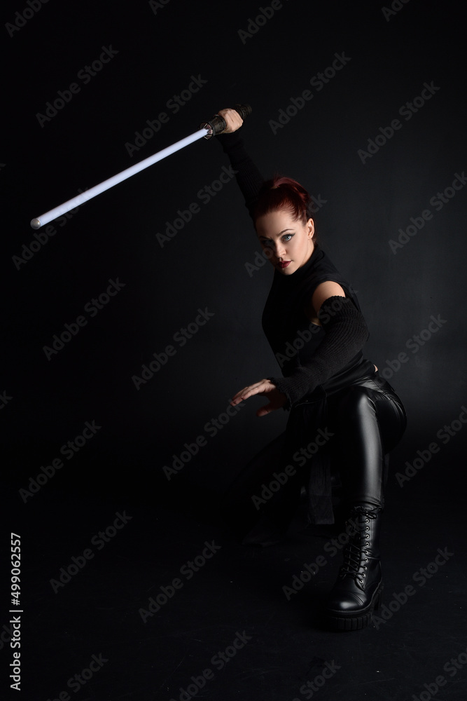 Full length portrait of pretty redhead female model wearing black futuristic scifi leather cloak costume. Standing pose holding lightsaber on dark studio background with shadow moody lighting. Full length portrait of pretty redhead female model wearing black futuristic scifi leather cloak costume. Standing pose holding lightsaber on dark studio background with shadow moody lighting.