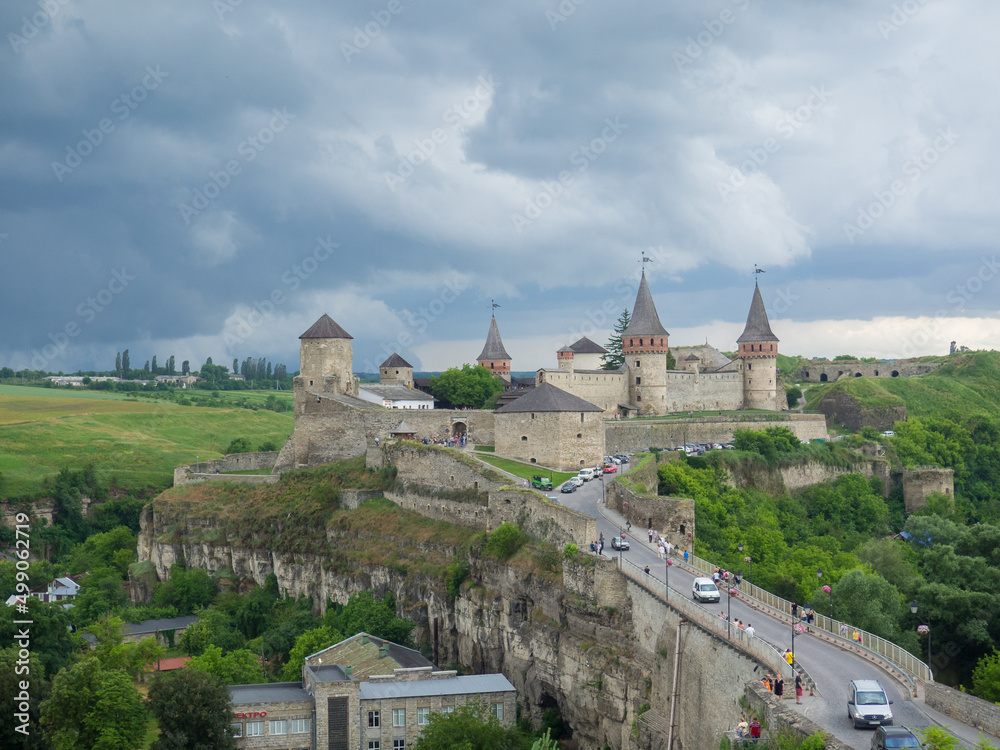 Kamianets Podilsky fortress on cloudy summer day. Scenic summer view of ancient fortress castle in Khmelnytskyi Region, Ukraine. Medieval stone large castle fortress with spiers and defensive towers. Kamianets Podilsky fortress on cloudy summer day. Scenic summer view of ancient fortress castle in Khmelnytskyi Region, Ukraine. Medieval stone large castle fortress with spiers and defensive towers.
