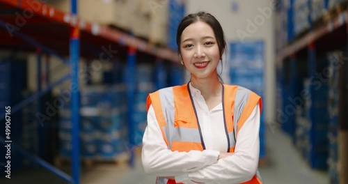 portrait of smiling Asian woman warehouse worker in protective vest standing with cross arm looking at camera in large warehouse