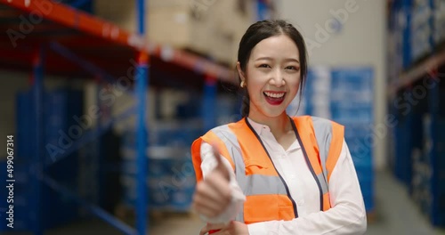 portrait of smiling Asian woman warehouse worker in protective vest standing with cross arm looking at camera in large warehouse