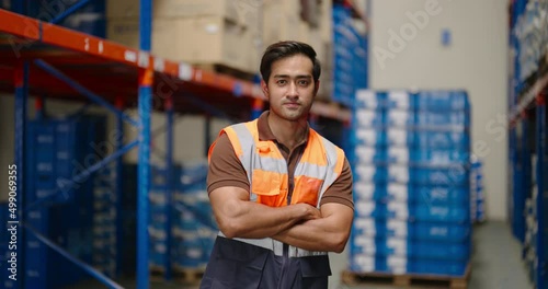 portrait of smiling Asian man warehouse worker in protective vest standing with cross arm looking at camera in large warehouse