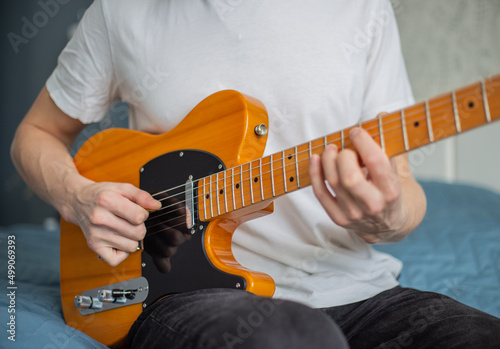 A man in a white T-shirt plays a natural-colored electric guitar sitting on a bed in close-up, selective focus.A male musician plays an electric guitar.Telecaster of natural color.Man playing guitar. 