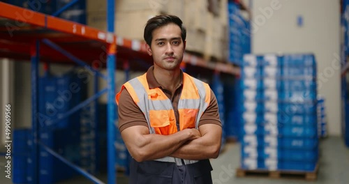 portrait of smiling Asian man warehouse worker in protective vest standing with cross arm looking at camera in large warehouse