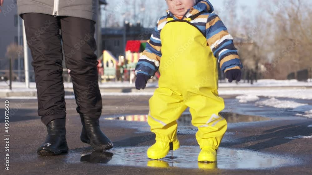 Mom with small cute child in yellow waterproof rain pants bibs for kids walking outdoors in cold season, one year old baby boy walking in a puddle. High quality 4k footage Mom with small cute child in yellow waterproof rain pants bibs for kids walking outdoors in cold season, one year old baby boy walking in a puddle. High quality 4k footage