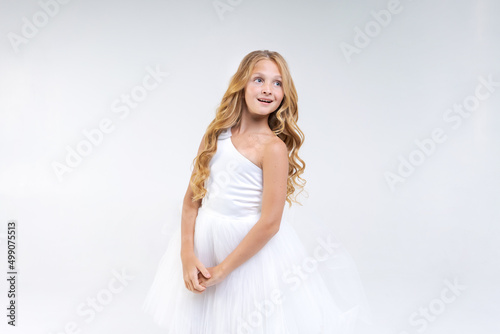 Beautiful cute girl in white dress with long wavy hair posing in studio on background. Caucasian school age girl model