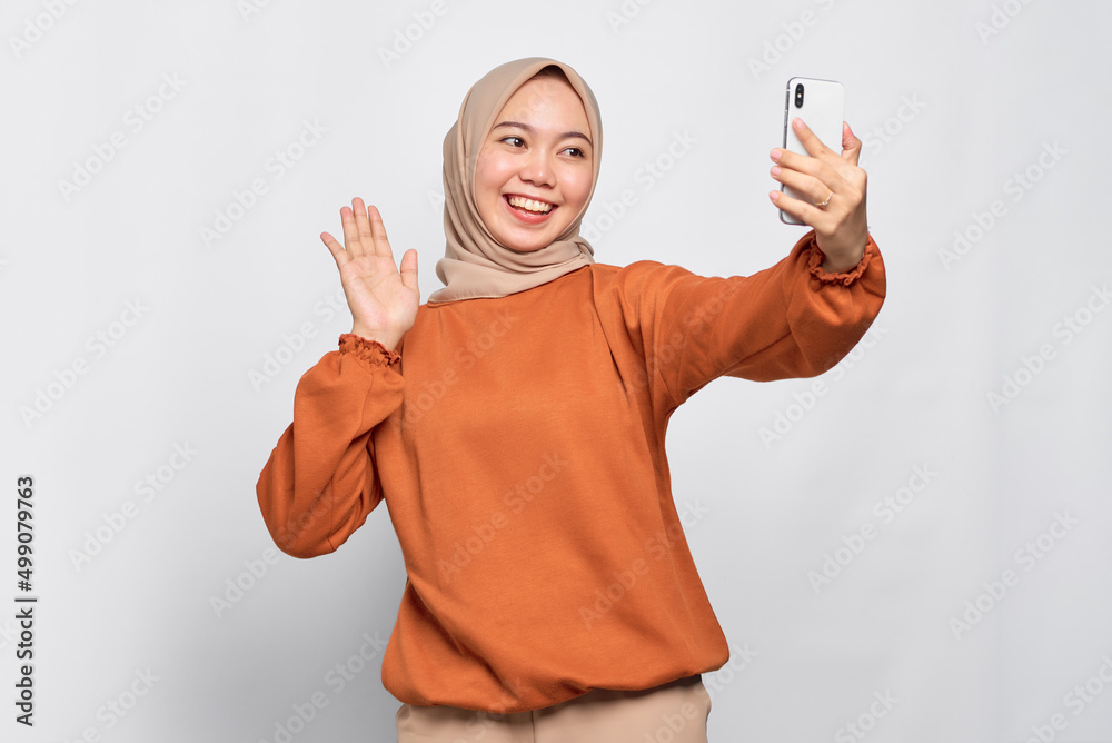 Cheerful young Asian woman in orange shirt doing selfie shot on mobile phone waving hand isolated over white background